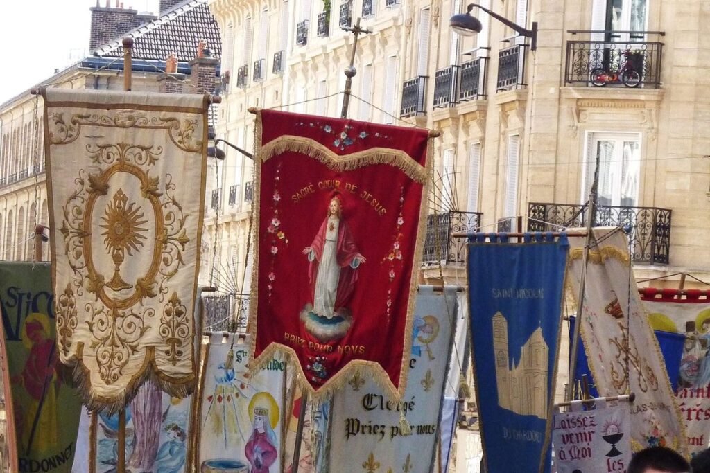 Bannières brodées de paroisses portées en procession dans une rue bordée d'immeubles en pierre, dont une bannière rouge du Sacré-Cœur