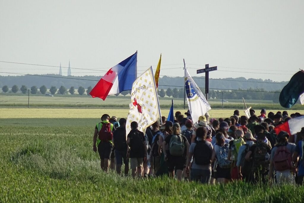 Groupe de pèlerins vus de dos, sacs au dos et drapeaux levés, marchant à travers les champs de la Beauce vers les flèches de la cathédrale de Chartres